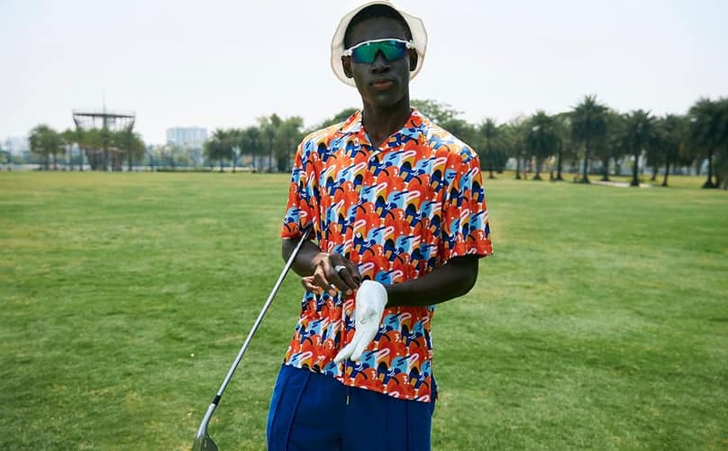 A golfer on the fairway in a NANANA orange-and-blue graphic print polo, bucket hat and sunglasses, holding a club.