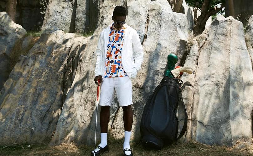 A golfer in a white jacket over a NANANA graphic polo, standing against rocks beside a golf bag.