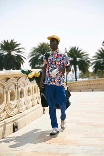 A golfer walking a palm-lined promenade carrying clubs, in a yellow cap and a red, blue and yellow geometric NANANA polo.